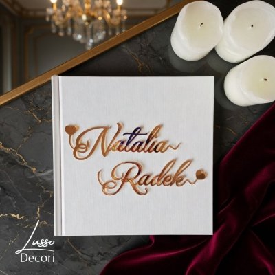 Biała kwadratowa księga gości z lustrzanymi złotymi imionami wykonanymi ozdobną czcionką.